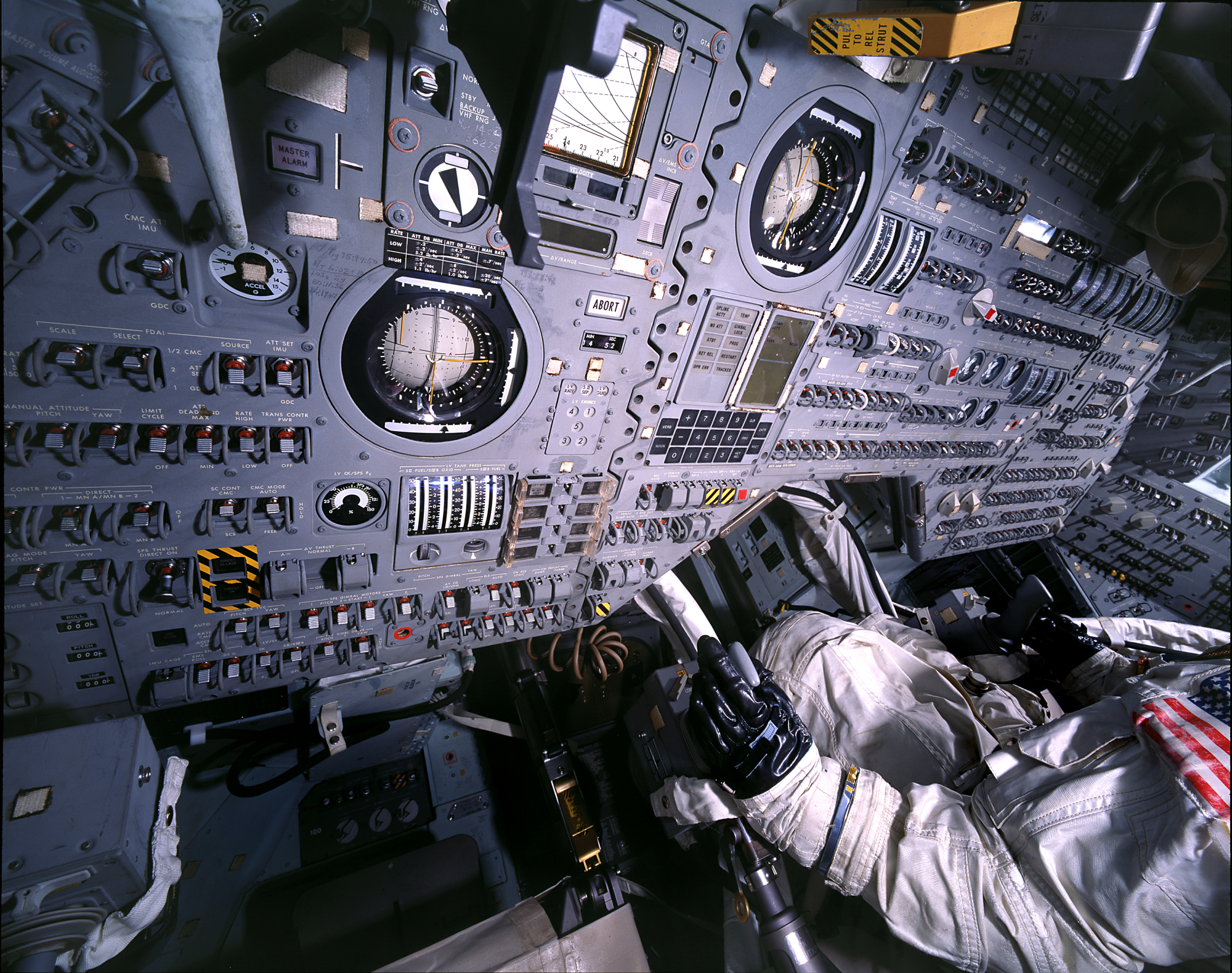 Close up of a control panel with innumerable buttons and switches. On the right mannequin of an astronaut sits, demonstrating the cramped conditions on board and the complexity of the spacecraft.
